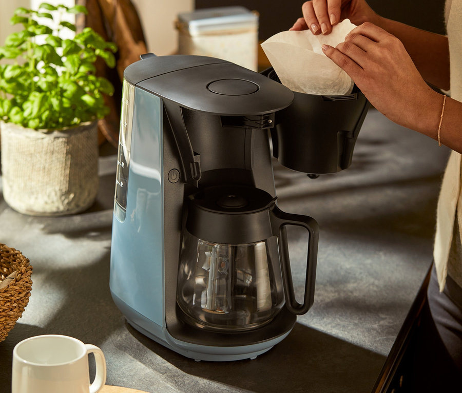 Frau legt einen Papierfilter in eine graublaue »Let's Brew« Filterkaffeemaschine ein.
