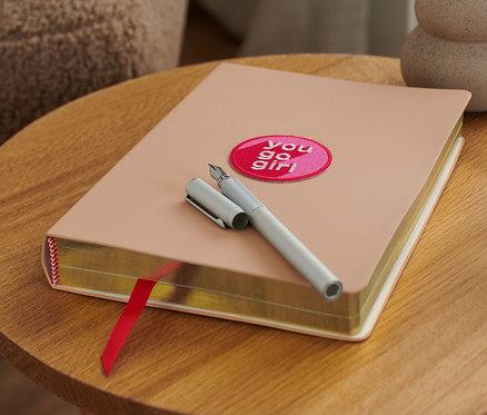 Beiges Notizbuch mit Textilpatch und silberner Stift liegen auf einem runden Holztisch neben einem Glas Wasser.