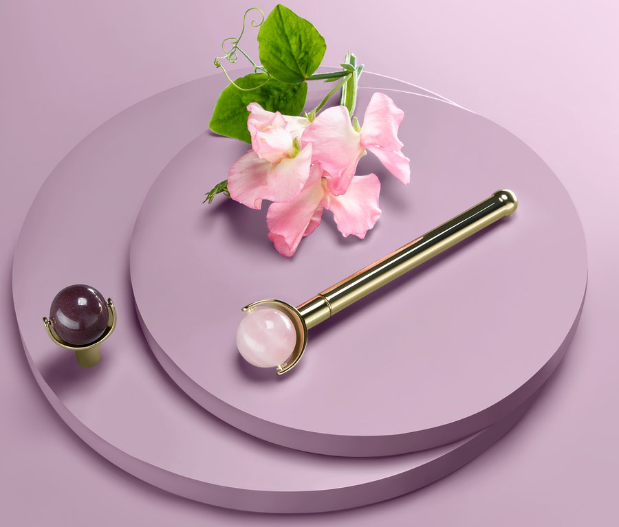 Ein Edelstein-Gesichtsroller liegt neben einer Blüte auf einem lila Hintergrund.
