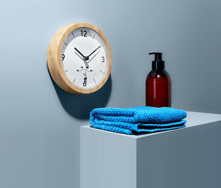 Eine Badwanduhr mit Thermometer hängt an der Wand. Auf einem Sockel liegen ein blaues Handtuch und eine braune Flasche mit Spender.