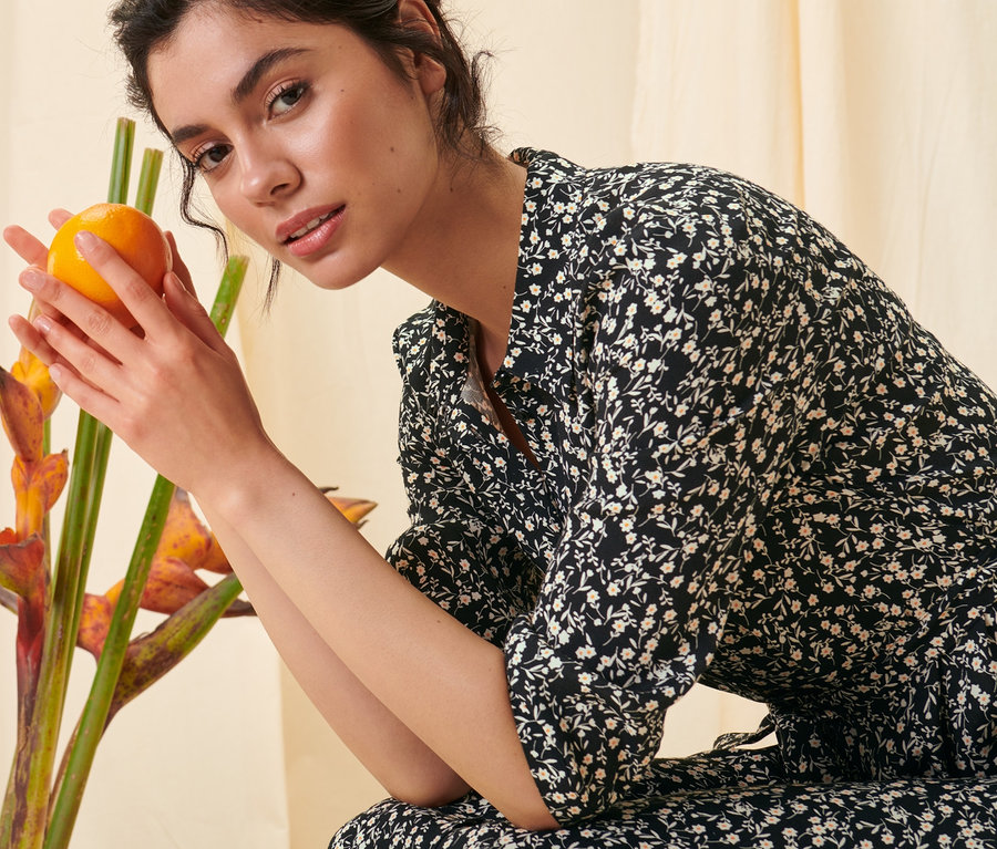 Frau hält Orange neben Blumen in einem floralen Hemdblusenkleid.