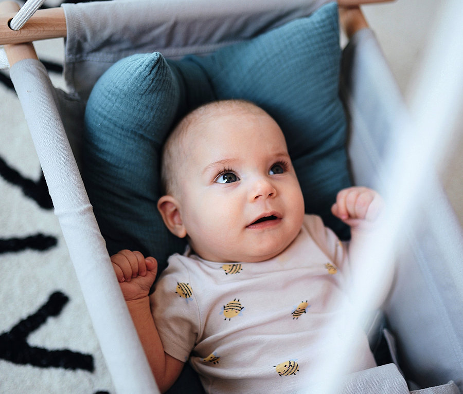 Ein Baby liegt in einer grauen small foot Babyschaukel »Seaside« mit blauem Kissen auf einer Unterlage mit schwarz-weißem Alphabet-Muster.