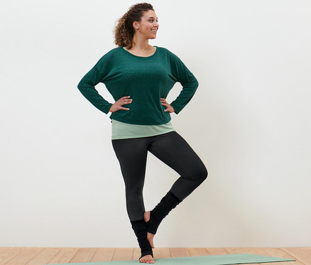 Frau steht in grünem 2-in-1-Sportshirt, Sporttight und schwarzen Stulpen auf einer Yogamatte.