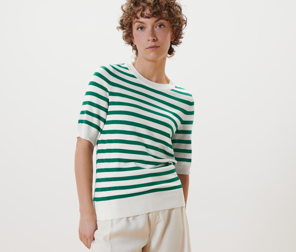 Frau posiert in einem gestreiften Feinstrickpullover, einer weißen Hose und weißen Schuhen.