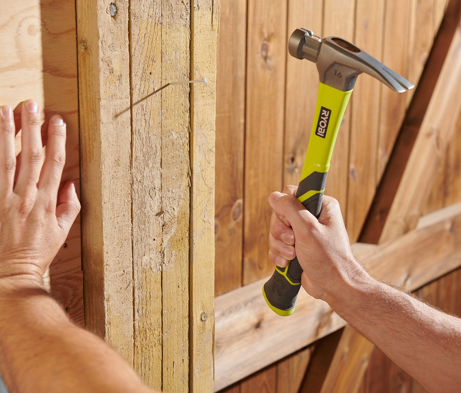 Mann hämmert mit einem Hammer einen Nagel in eine Holzwand. Er benutzt ein RYOBI 18 V ONE+ Heimwerker-Set »R18PD3-220TAH«.