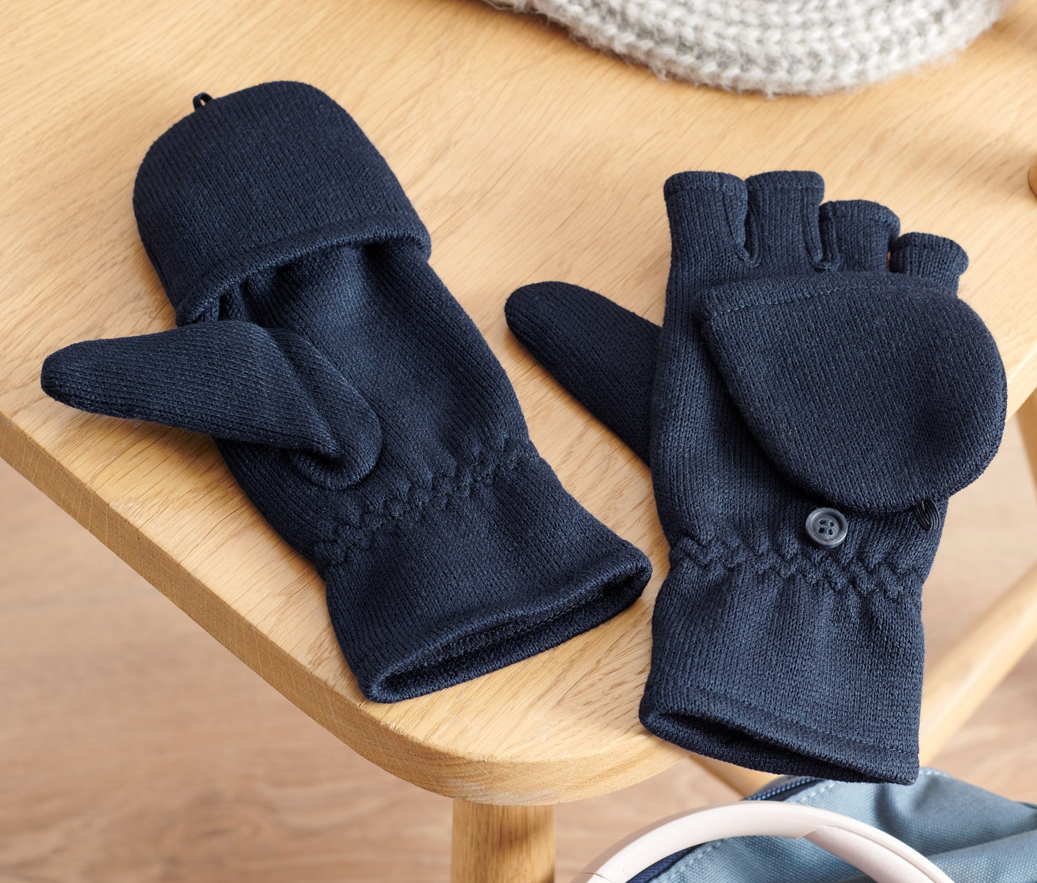 Kinder-Strickfleece-Klappfäustlinge, eine Strickmütze und Kopfhörer liegen auf einem Holzstuhl auf einem Rucksack.