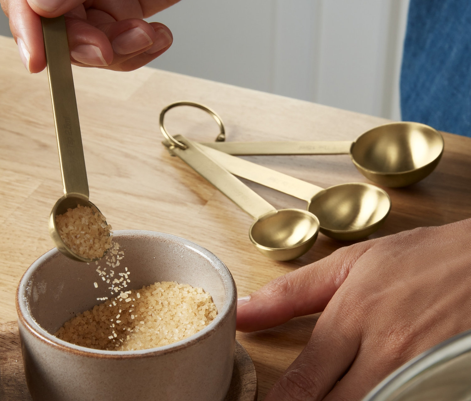 Eine Hand giesst mit einem Messlöffel braunen Zucker in eine Schüssel. Auf dem Tisch liegt ein Messlöffel-Set.