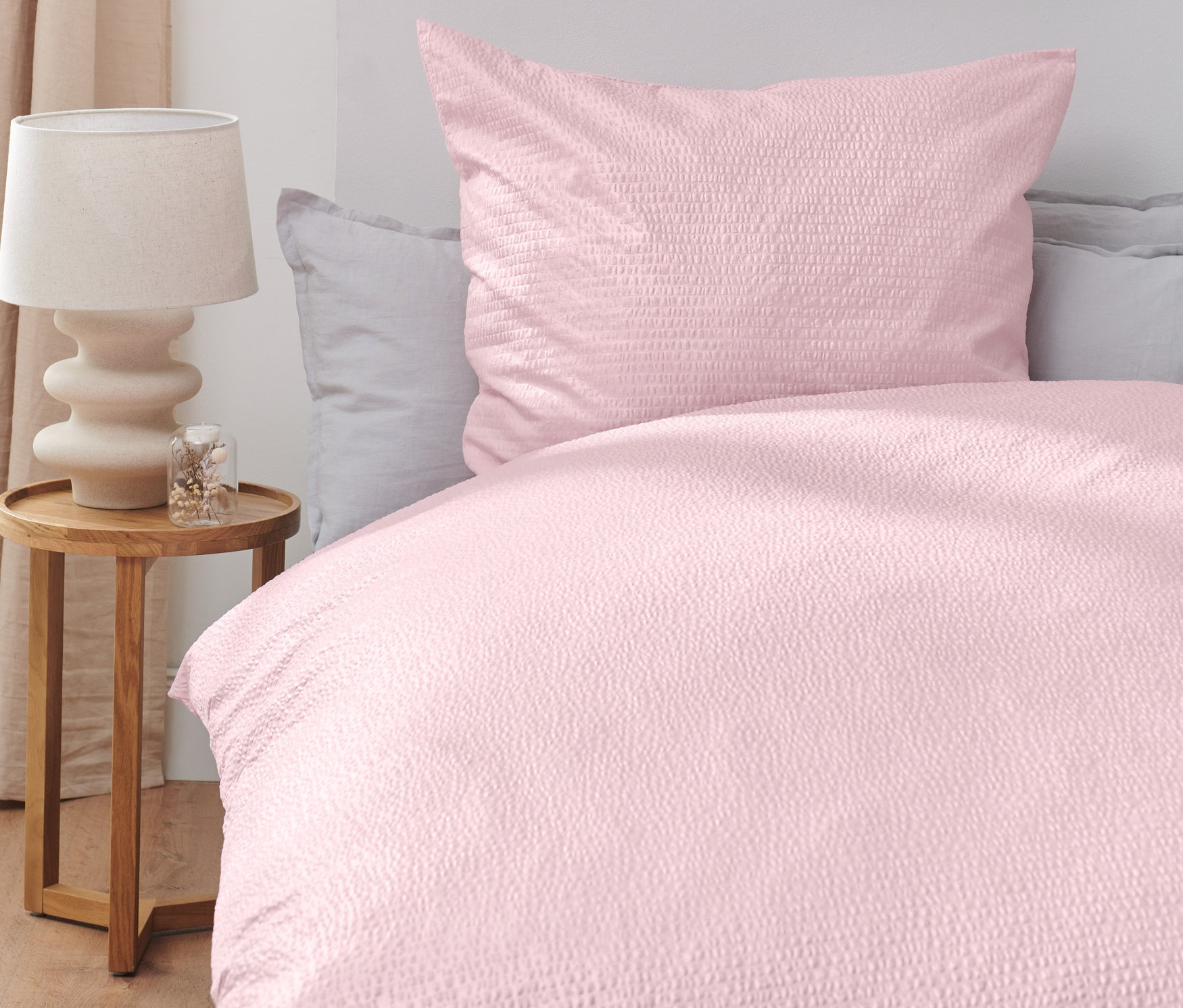 Rosa Seersucker-Bettwäsche auf einem Bett, daneben ein Holztisch mit Lampe.