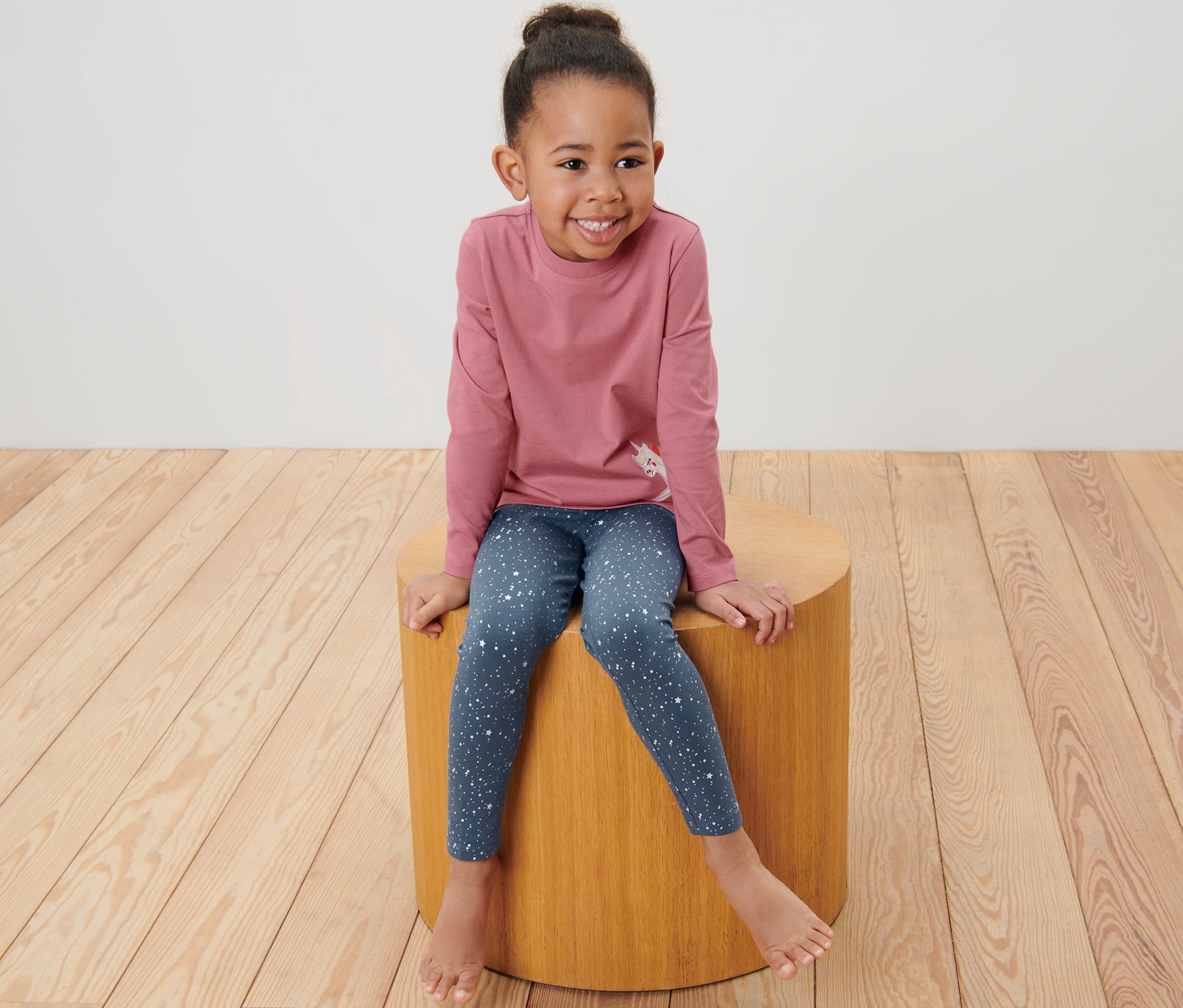 Ein lächelndes Mädchen sitzt auf einem Holzhocker und trägt 2 Kinder-Glitzer-Leggings.