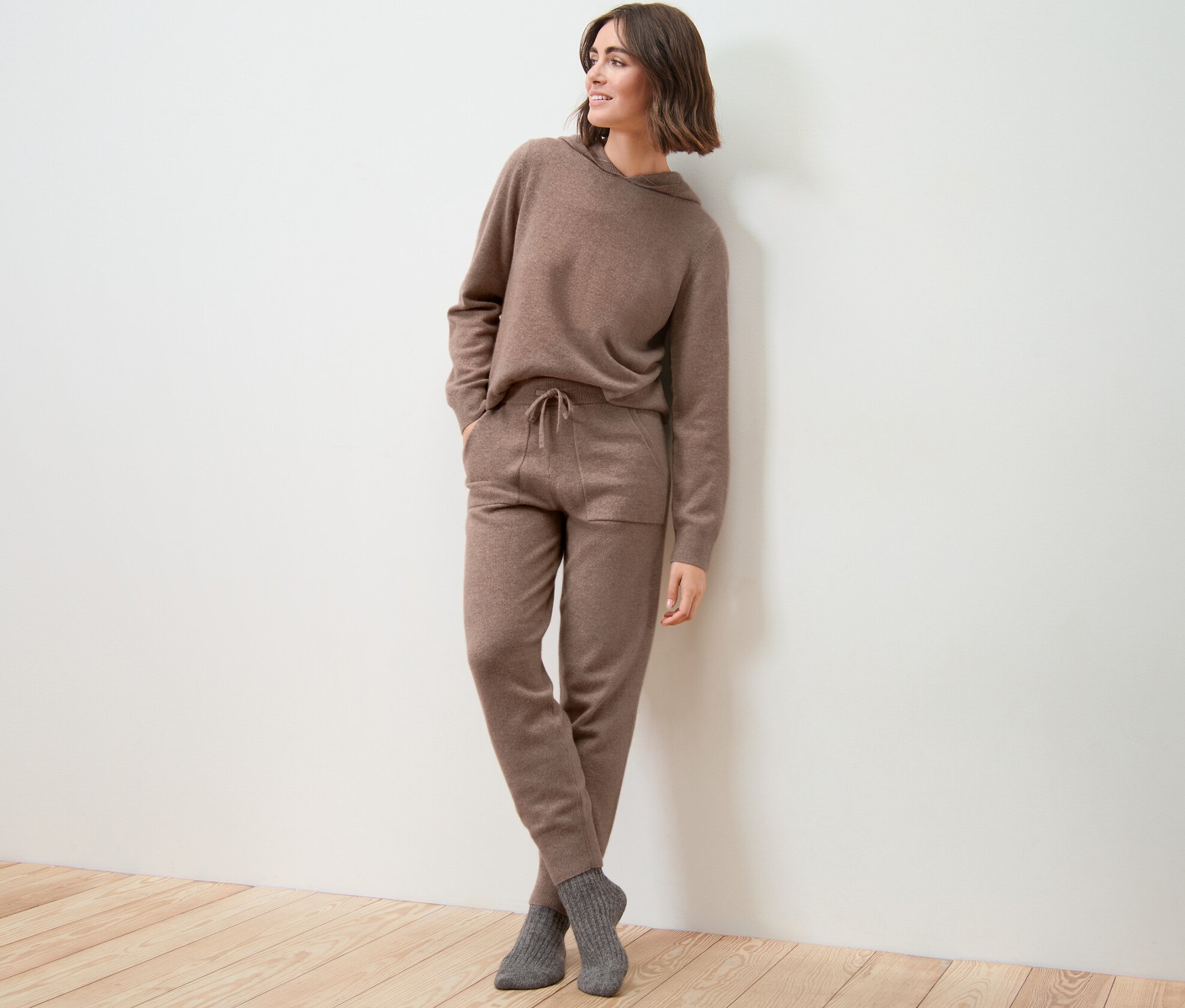 Frau in brauner Cashmere-Loungewearhose und Kapuzenpullover steht an einer Wand.