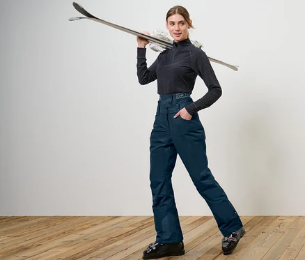 Frau posiert in marineblauer Ski- und Snowboardhose und hält Ski über der Schulter.