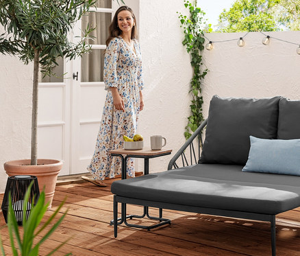 Eine Frau im Kleid steht neben einem C-Table aus Aluminium und einem ausziehbaren Lounge-Sofa. Im Hintergrund befindet sich eine Solar-Lichterkette, Kugeln mit Rillen.
