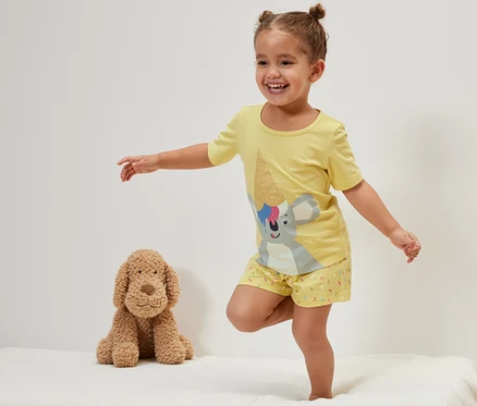 Ein lächelndes kleines Mädchen steht auf einem Bett neben einem Plüschhund und trägt einen gelben Kinder-Shorty-Pyjama, Koala.