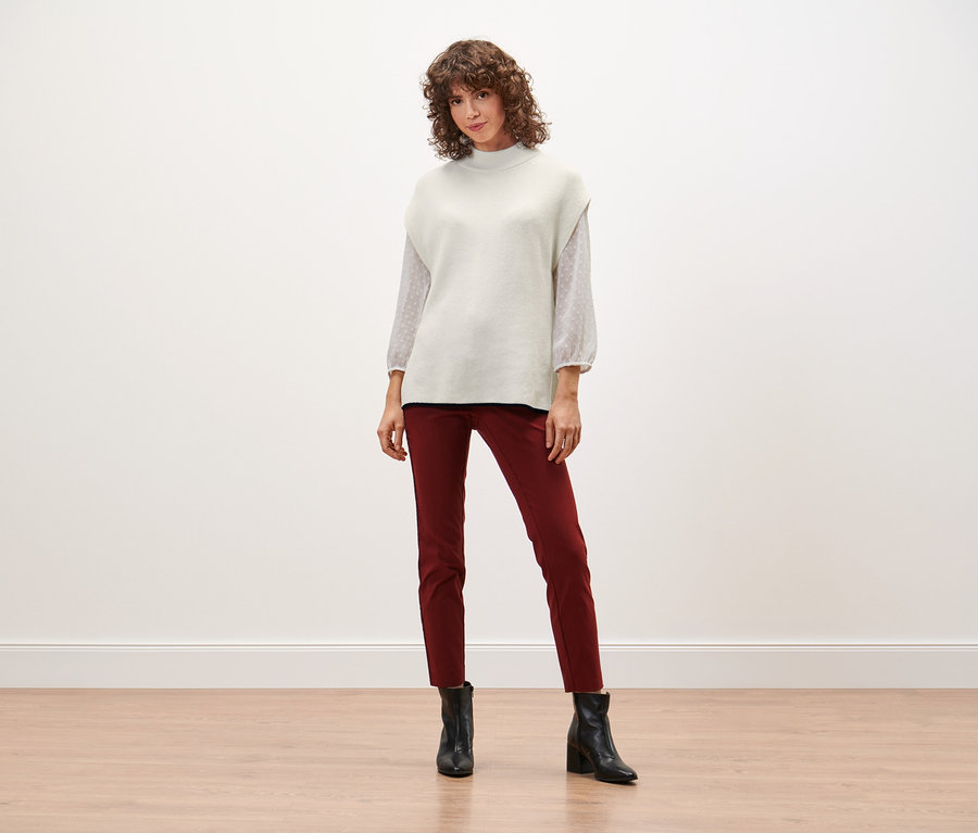 Frau steht und trägt einen Strick-Pullunder, rote Hosen und schwarze Stiefel.