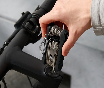 Eine Hand hält ein Fahrrad-Multifunktionswerkzeug am Lenker eines Fahrrads. Im Hintergrund ist ein Helm zu sehen.
