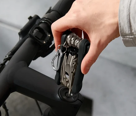 Eine Hand hält ein Fahrrad-Multifunktionswerkzeug am Lenker eines Fahrrads. Im Hintergrund ist ein Helm zu sehen.