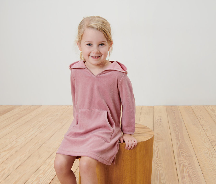 Ein lächelndes Mädchen sitzt auf einem Holzhocker und trägt einen Kinder-Frottierponcho.