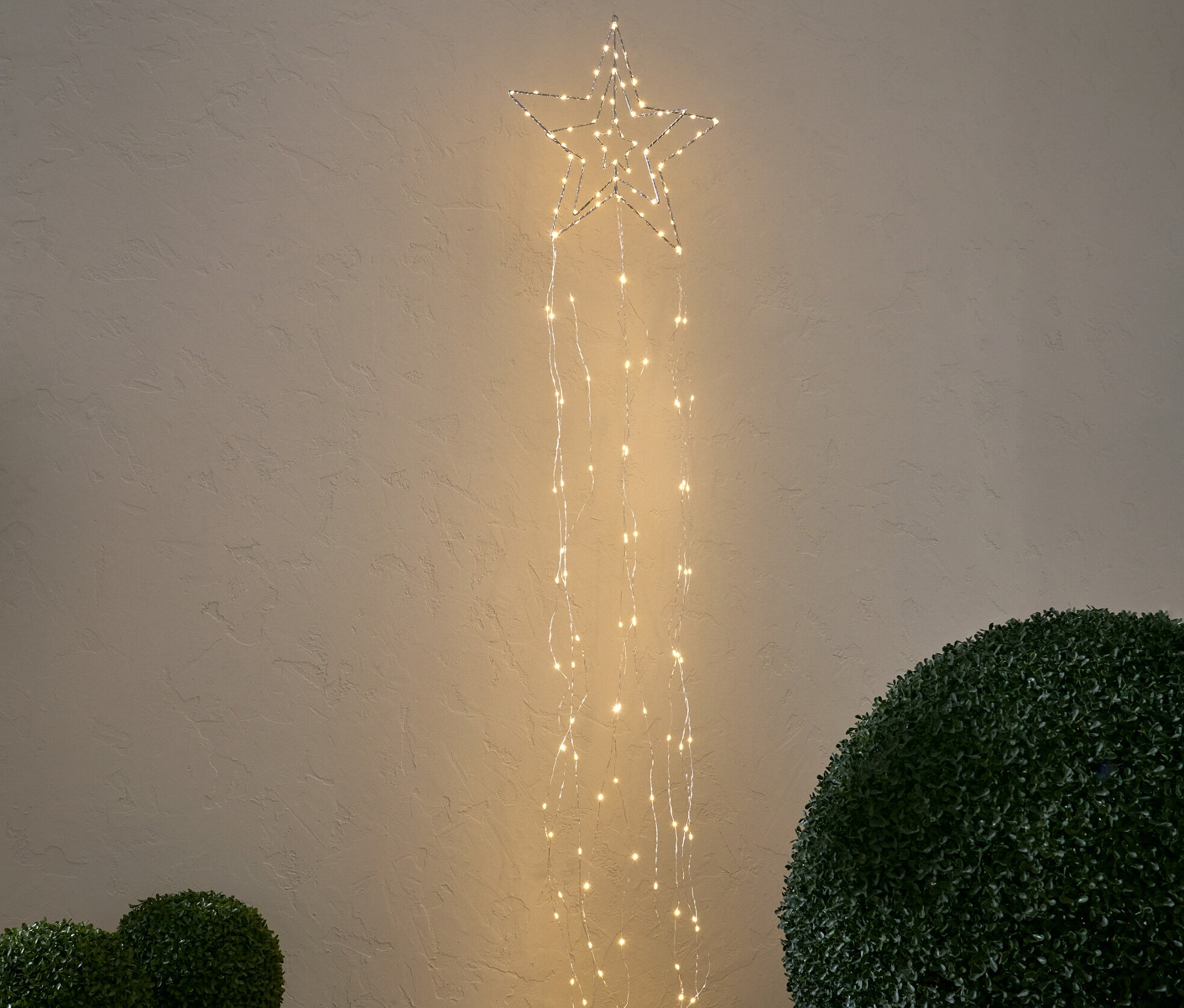 Ein LED-Stern mit Lichterbündel hängt vor einer Wand. Darunter stehen zwei kleine Büsche in Töpfen und ein großer.