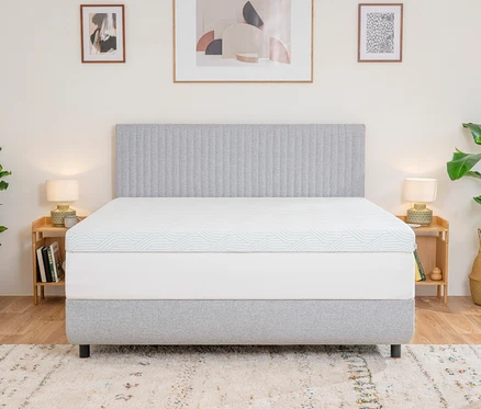 Ein Bett mit grauem Kopfteil und Untergestell, darauf ein weißer TEMPUR PRO® Plus SmartCool™ Topper mit blauen Wellenlinien. Holznachttische mit Lampen und Pflanzen flankieren das Bett, ein gemusterter Teppich liegt auf dem Boden.