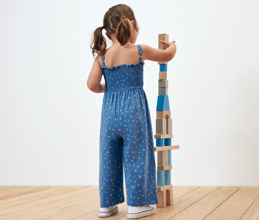 Mädchen baut Turm aus Holzklötzen. Sie trägt einen Jumpsuit in 7/8-Länge.