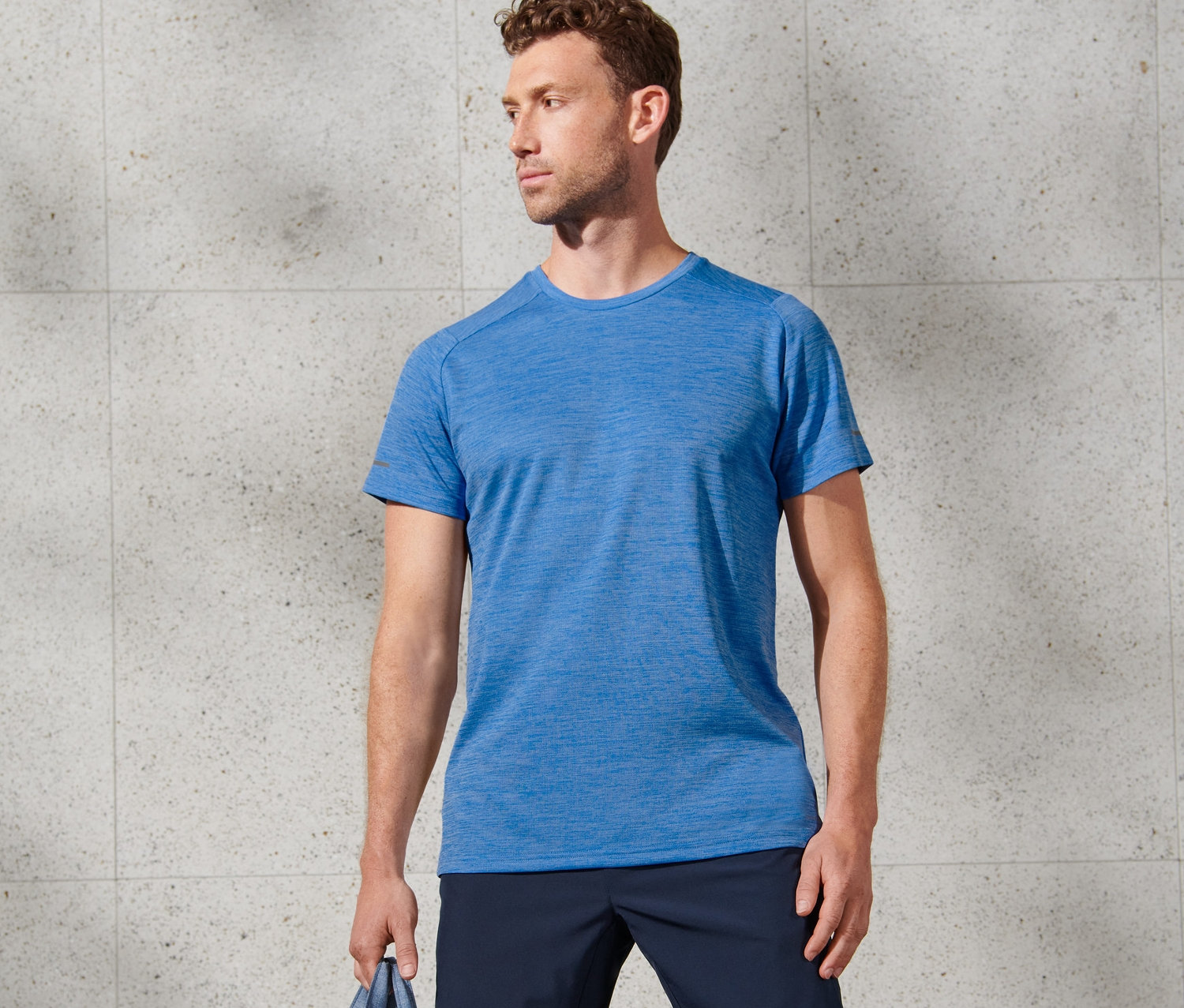 Mann posiert mit blau meliertem Funktionsshirt, dunkelblauen Shorts und Sporttasche.