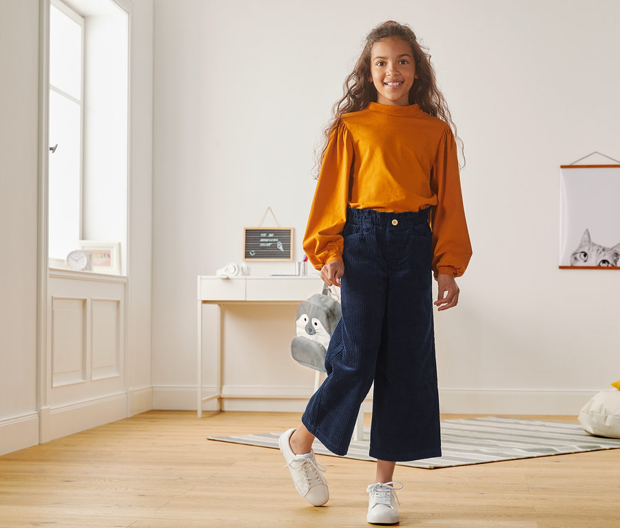 Mädchen posiert in orangefarbenem Oberteil und Cord-Culotte.