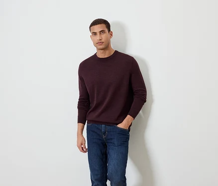 Mann posiert vor einem weißen Hintergrund und trägt einen burgunderfarbenen Pullover und Jeans.