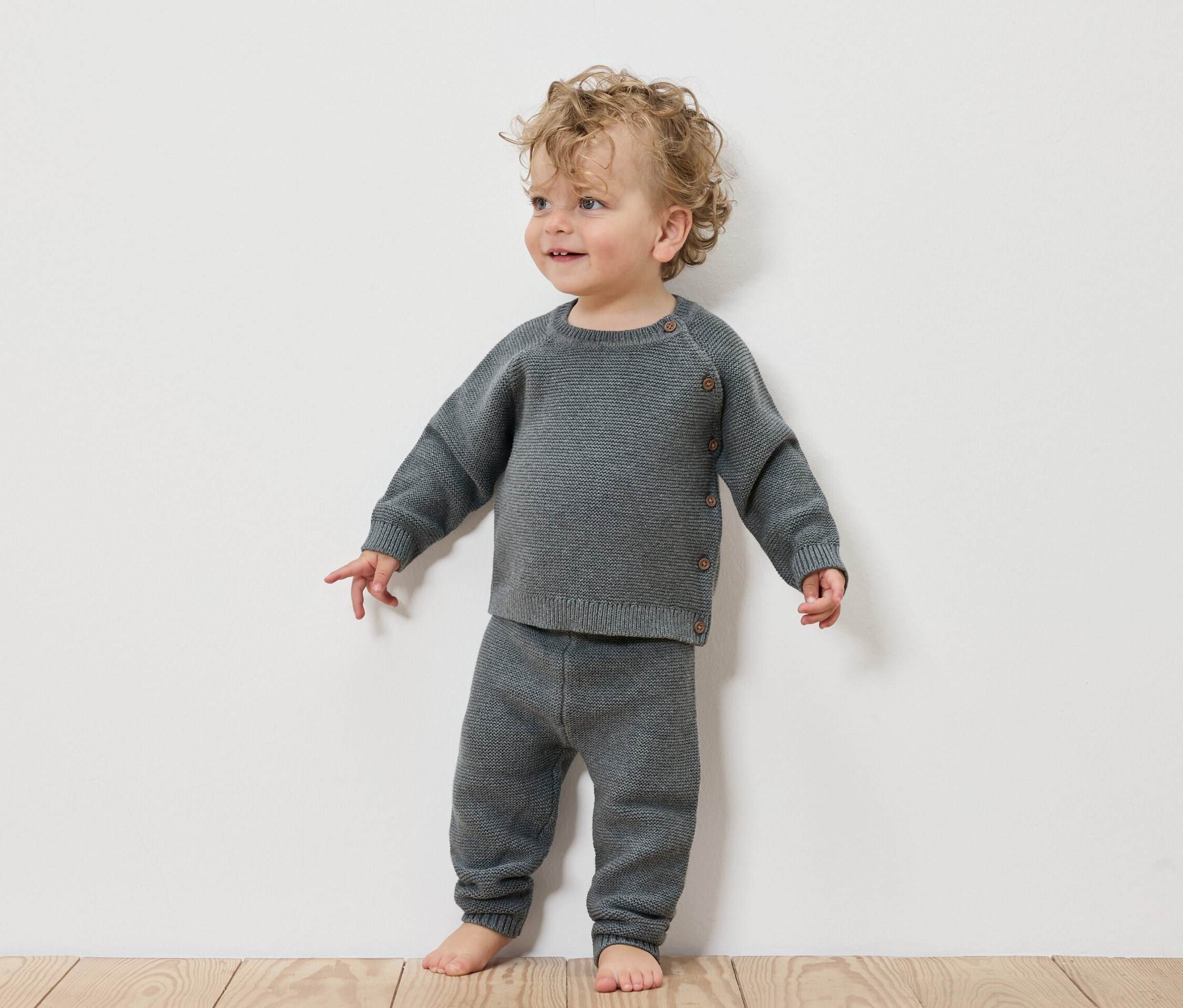 Kleinkind steht vor einer weißen Wand und trägt eine Baby-Strickkombi.