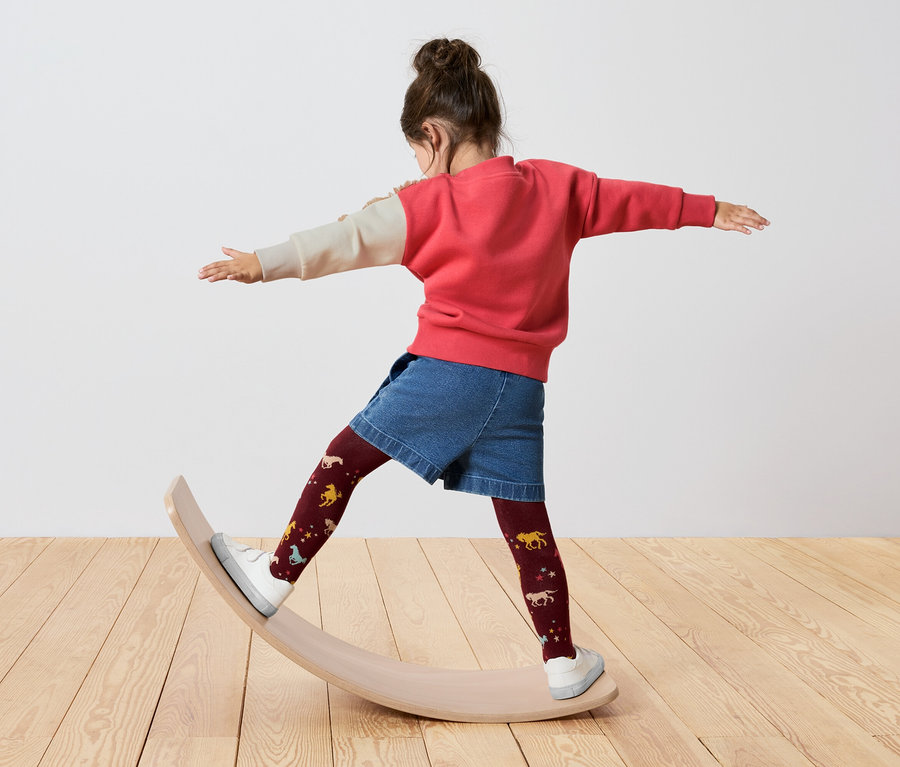 Mädchen steht auf einem Balance Board, trägt einen rot-cremefarbenen Pullover, Jeansshorts und bordeauxfarbene Strumpfhosen mit Pferden.