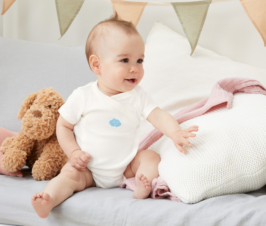 Baby sitzt auf einem Bett und trägt ein 5er-Pack Kurzarm-Bodys aus Bio-Baumwolle.
