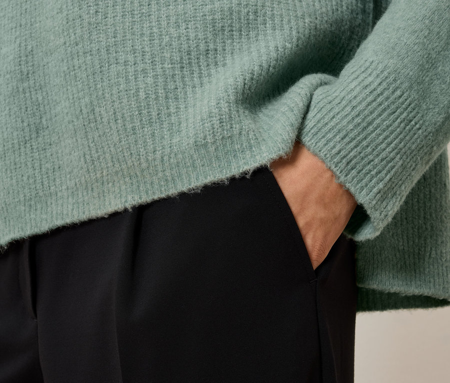Detailaufnahme eines Damenoutfits: Ein hellgrüner Pullover und eine schwarze Hose. Eine Hand steckt in der Hosentasche.
