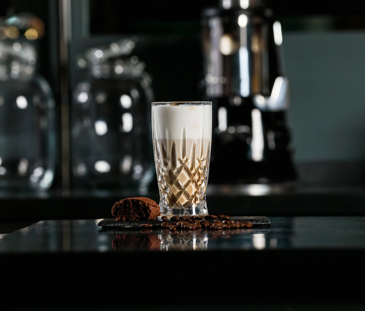 Ein Latte-Macchiato-Glas »Noblesse« von Nachtmann steht neben Kaffeebohnen und einem Keks auf einer glänzenden Oberfläche.