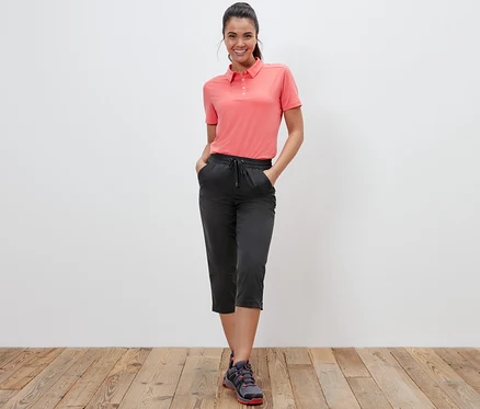 Eine Frau steht und posiert in einem korallenroten Polo-Shirt und einer Softshell-Funktionscapri.