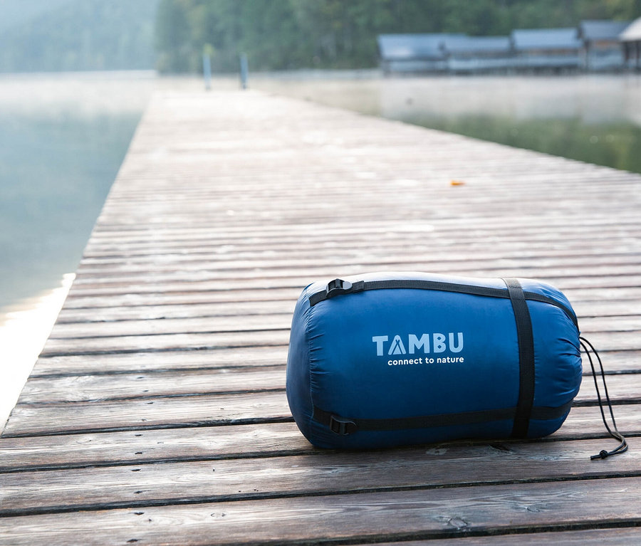 Blauer Tambu Mumienschlafsack Talu 1500 liegt auf einem Holzsteg an einem See.