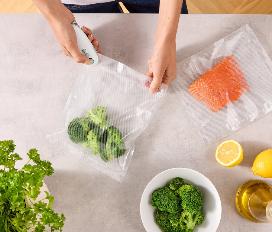 Eine Person verschließt mit einem Vakuumierer einen Beutel mit Brokkoli. Auf dem Tisch liegen ein weiterer Beutel mit Lachs, Zitronen, eine Schüssel Brokkoli, Petersilie und eine Ölflasche.
