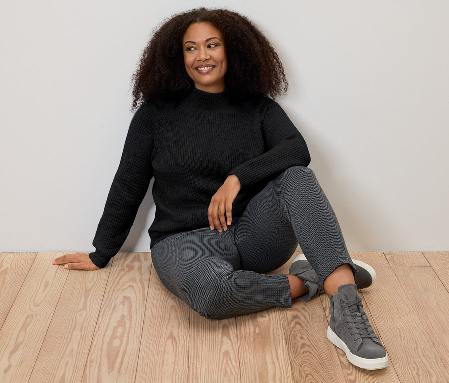 Frau sitzt auf dem Boden und trägt einen schwarzen Grobstrickpullover mit Stehkragen.