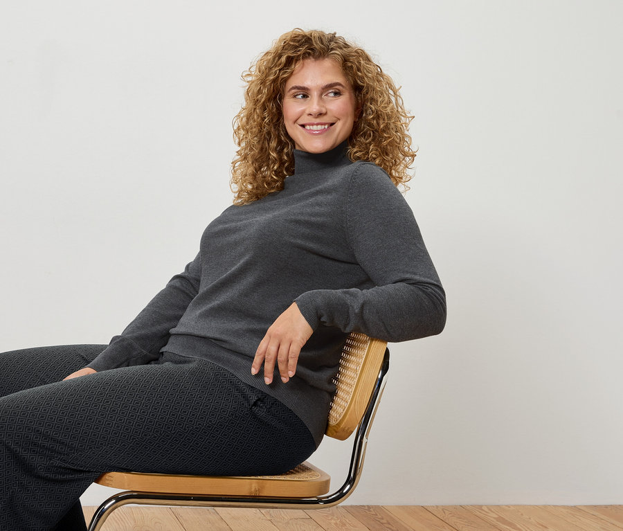 Eine Frau mit lockigem Haar sitzt auf einem Stuhl und trägt einen grauen Rollkragenpullover und gemusterte Hosen.