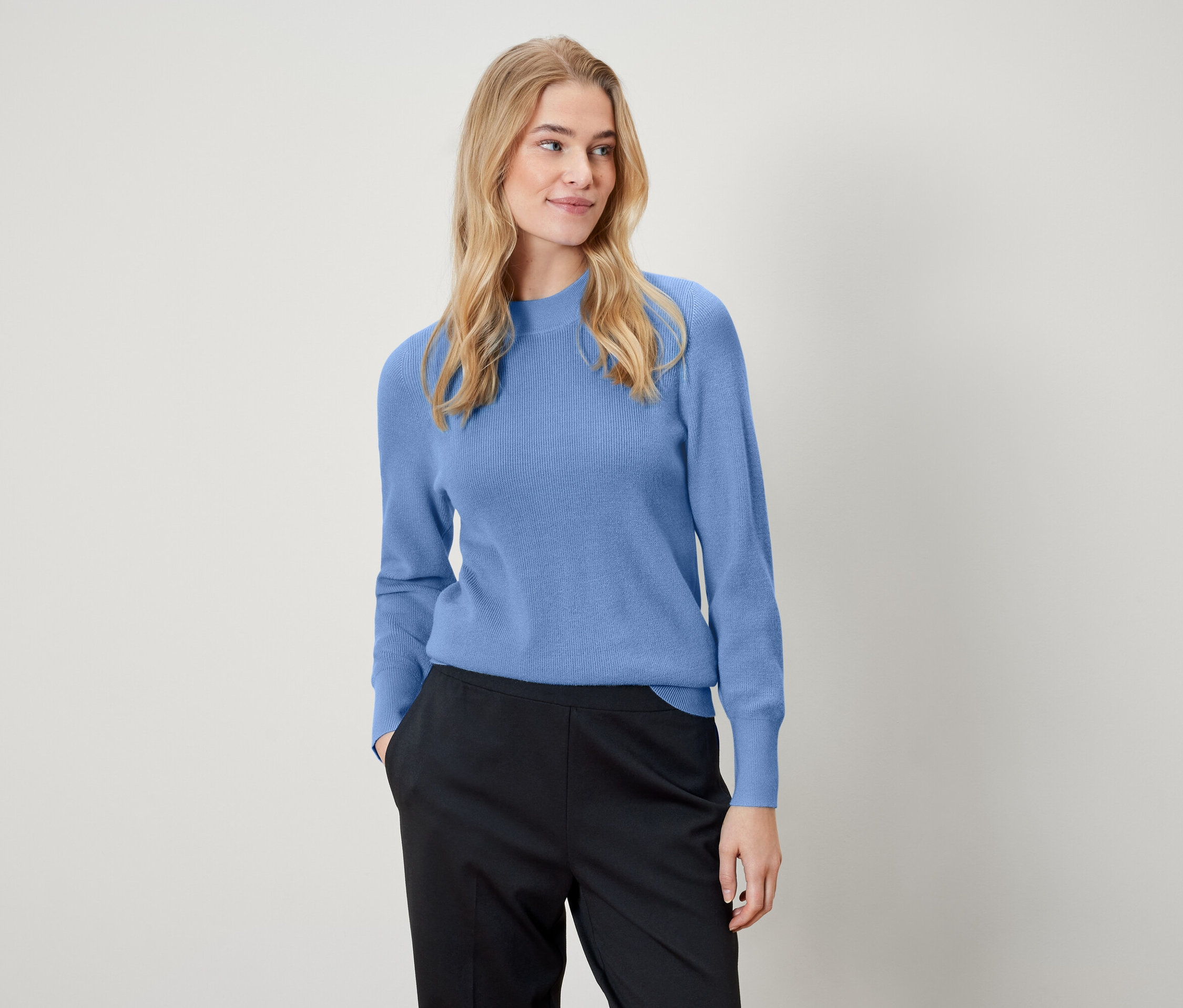 Blonde Frau trägt einen hellblauen Pullover und eine schwarze Hose.