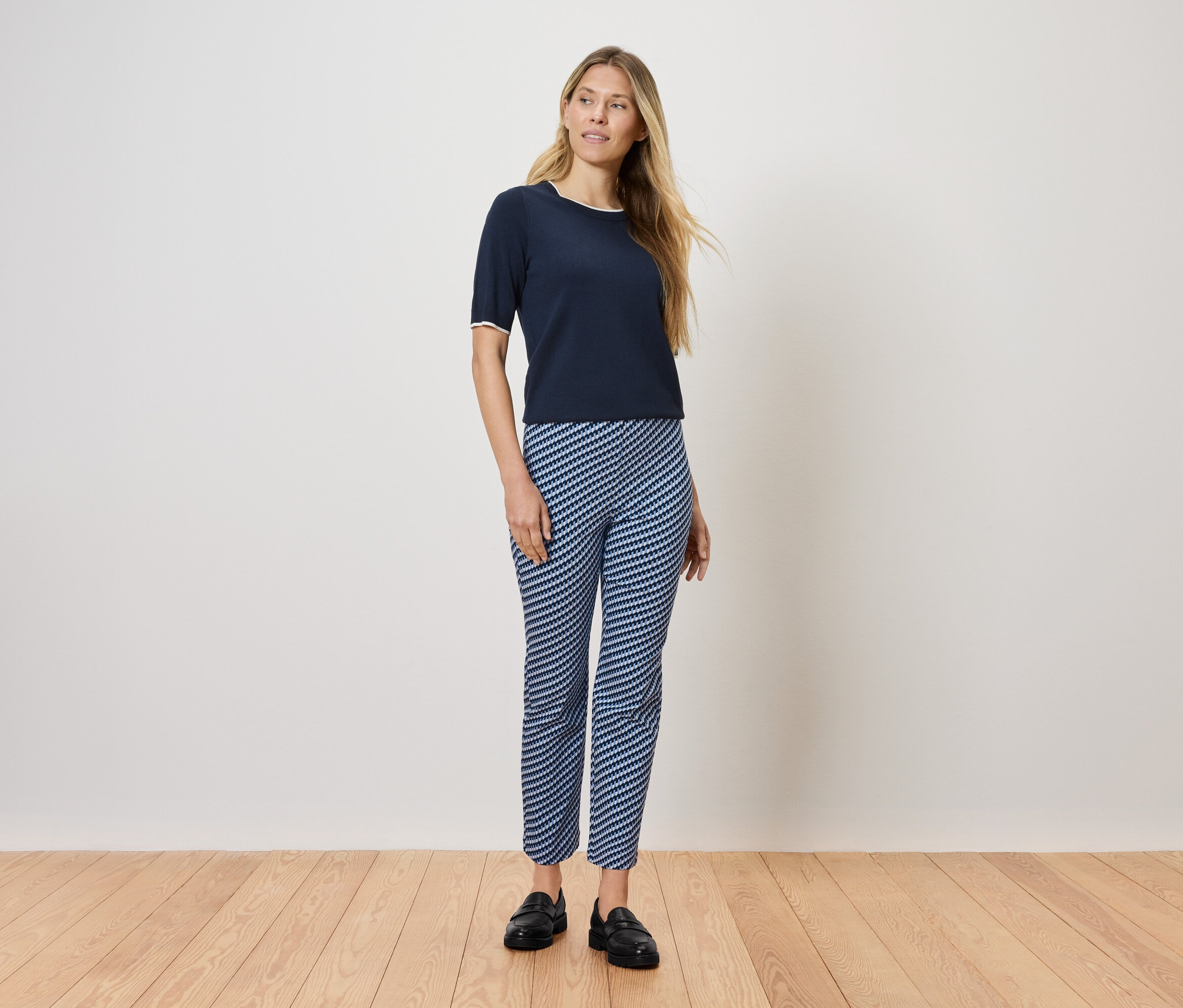 Eine Frau steht auf einem Holzboden und trägt ein dunkelblaues T-Shirt und blau-weiß gestreifte Hosen.