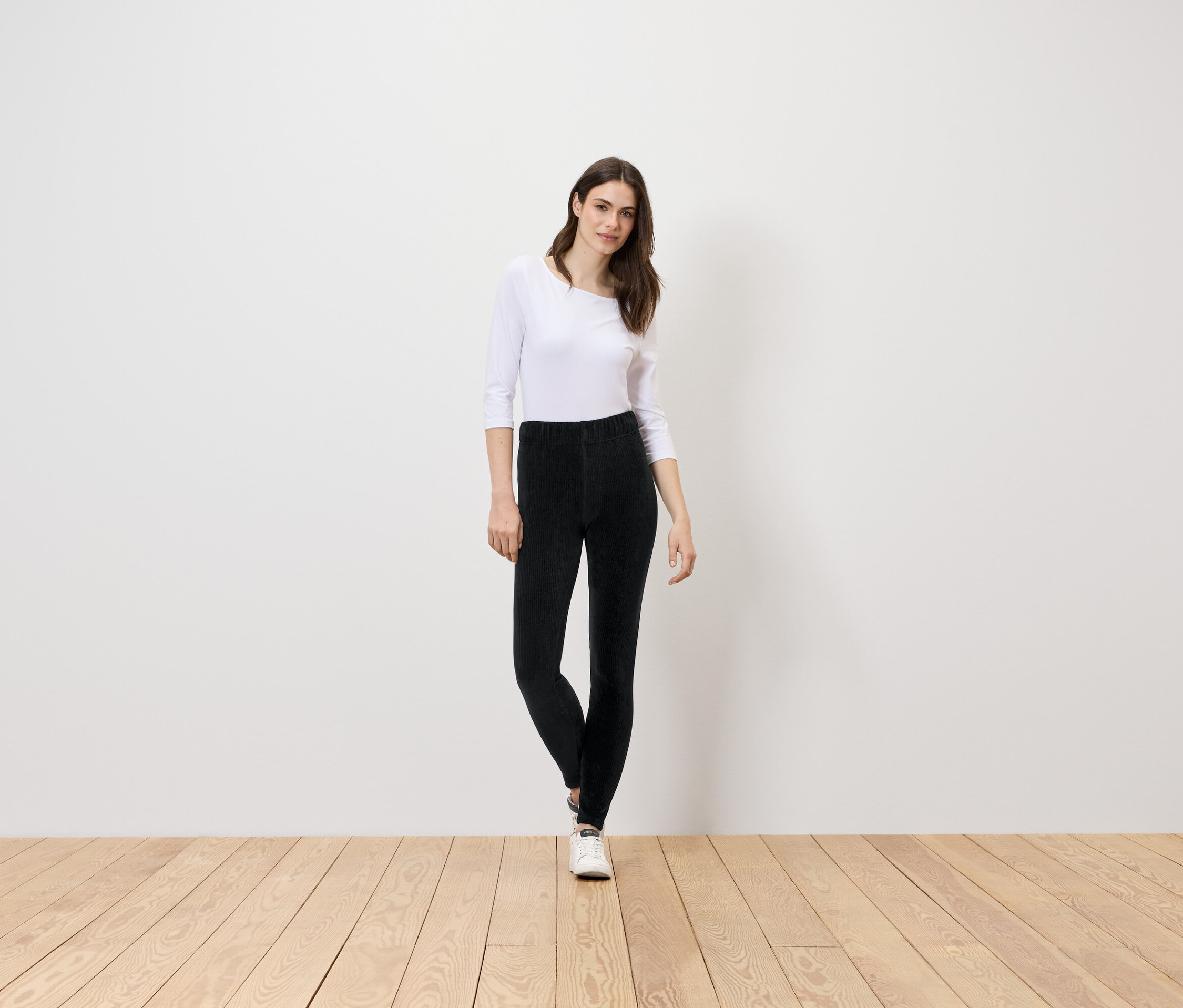 Frau steht auf Holzboden, trägt eine weiße Bluse und eine schwarze Hose.