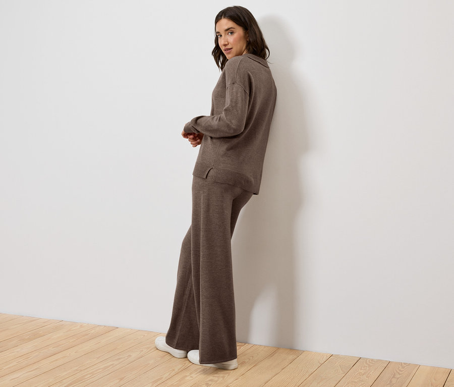 Eine Frau, die sich an eine Wand lehnt, trägt ein braunes Strickoberteil und eine Hose.