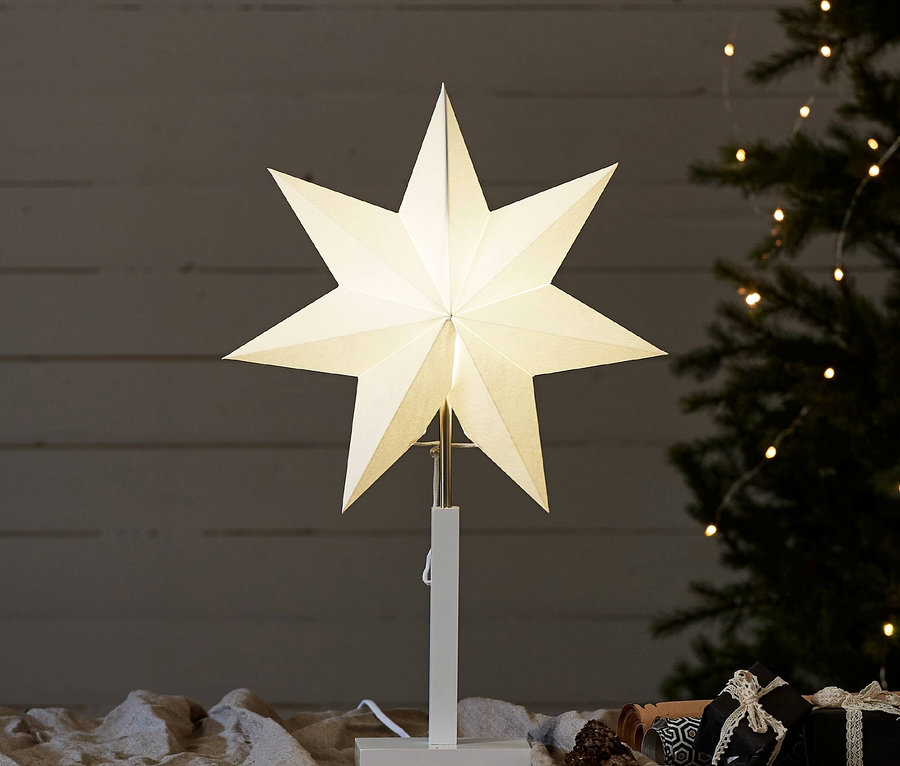 Ein leuchtender, weißer Stern steht auf einem Tisch, im Hintergrund ein Weihnachtsbaum.