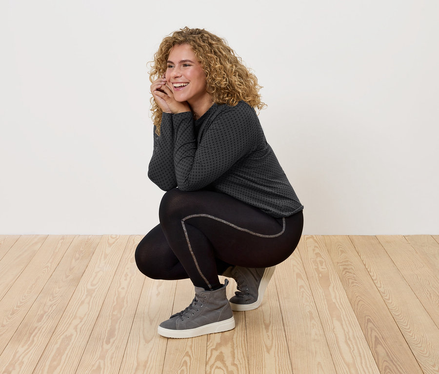 Frau mit lockigem Haar in schwarzer Sportbekleidung und grauen Turnschuhen posiert auf Holzboden.