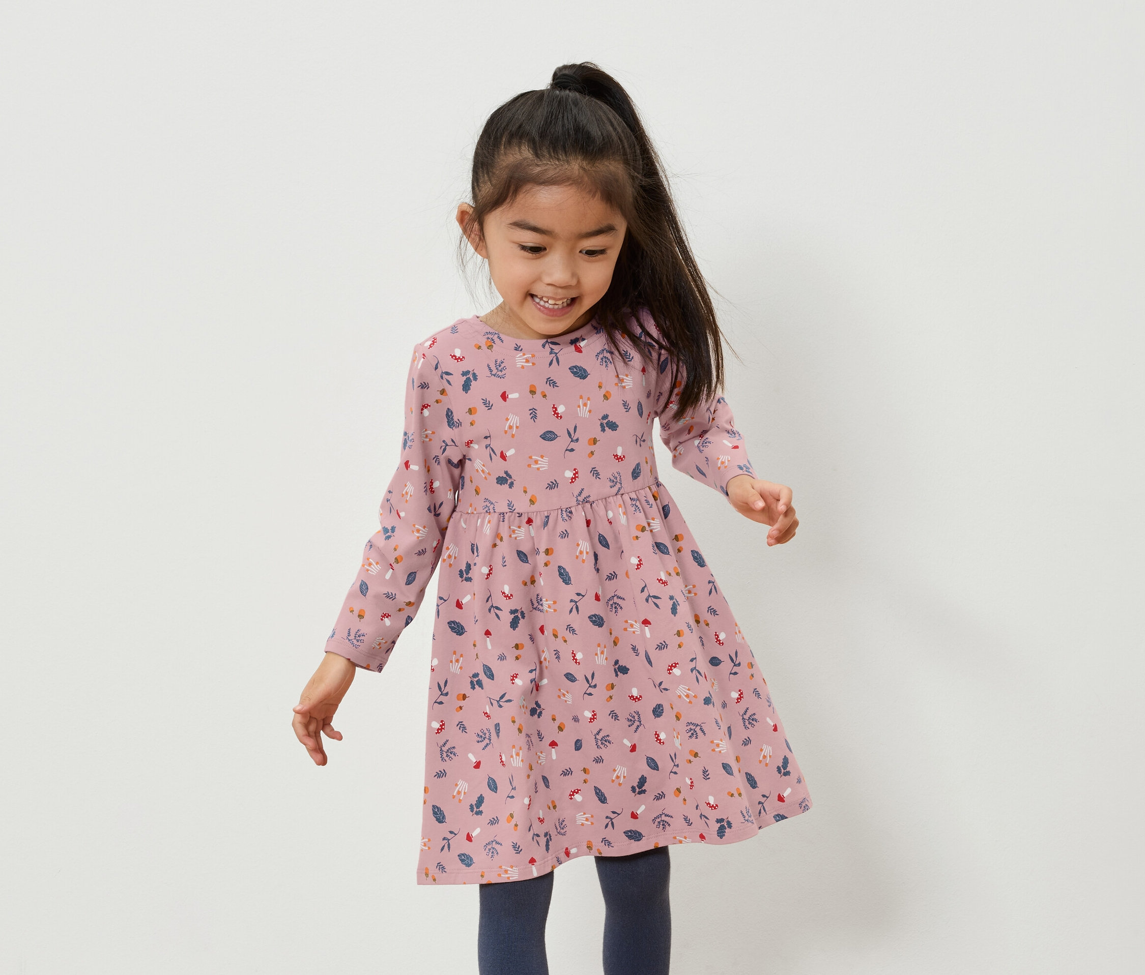 Lächelndes asiatisches Mädchen in einem rosafarbenen Kleid mit Blumenmuster und blauen Leggings.