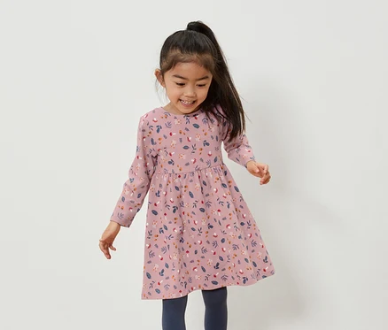 Lächelndes asiatisches Mädchen in einem rosafarbenen Kleid mit Blumenmuster und blauen Leggings.