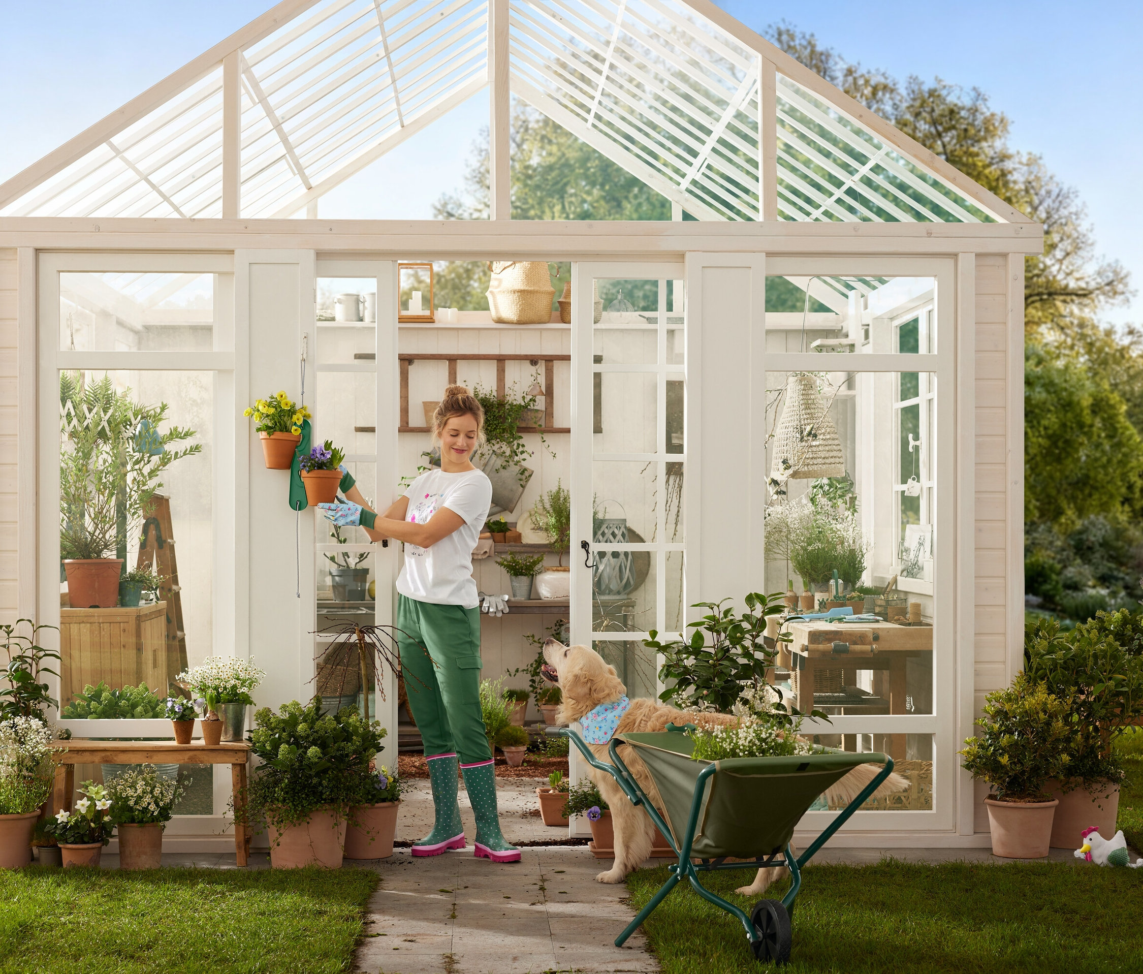 Eine Frau mit Gartenhandschuhen hängt Blumentöpfe an ein Gewächshaus, während ein Hund neben einer Schubkarre steht.