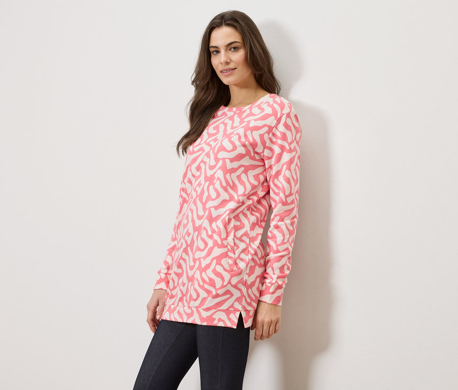 Frau trägt einen rosafarbenen Pullover mit abstraktem Muster und dunkle Jeans vor einem weißen Hintergrund.