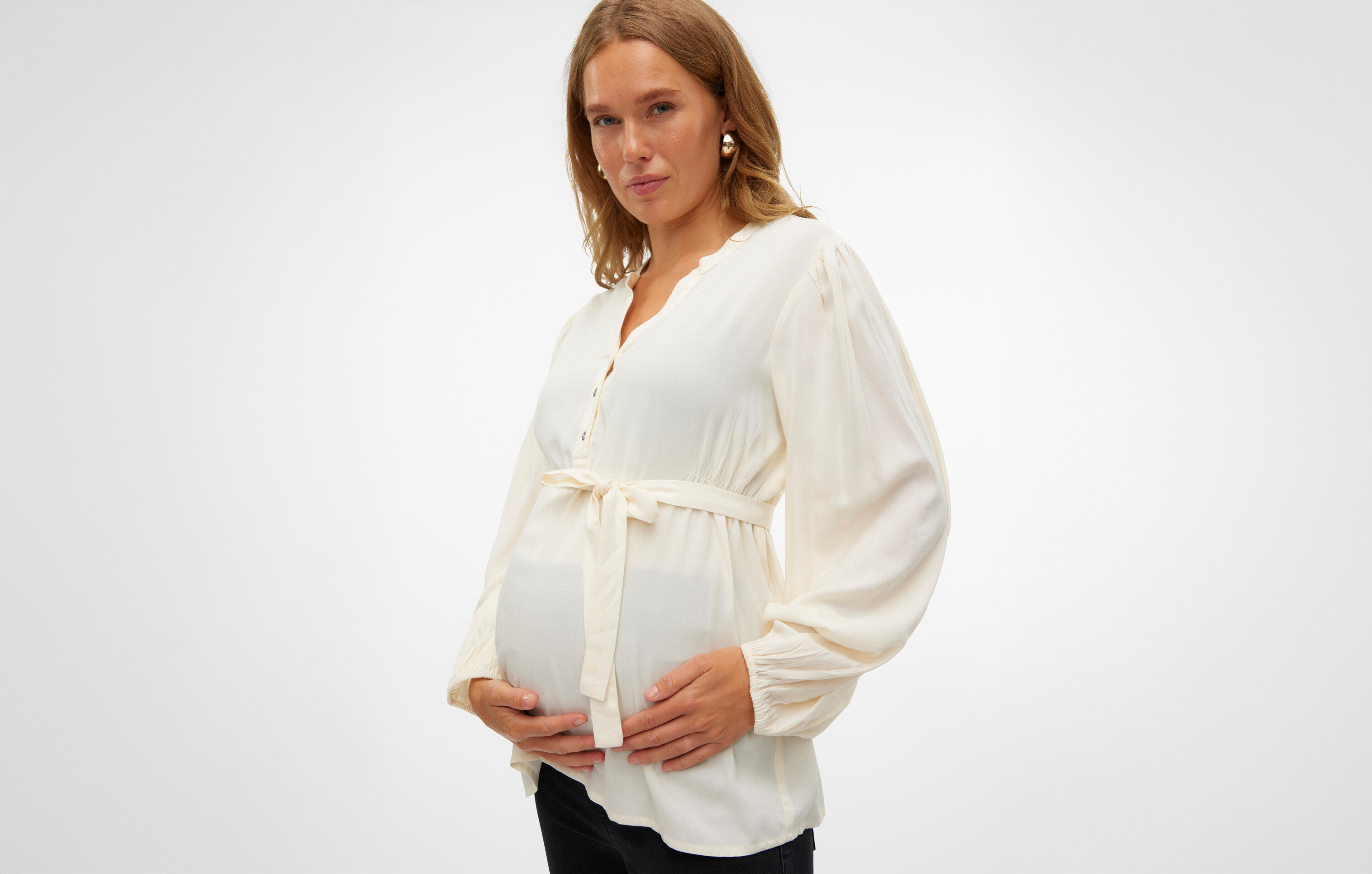 Schwangere Frau in einer weißen Bluse mit Gürtel hält ihren Bauch.