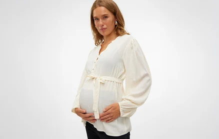 Schwangere Frau in einer weißen Bluse mit Gürtel hält ihren Bauch.