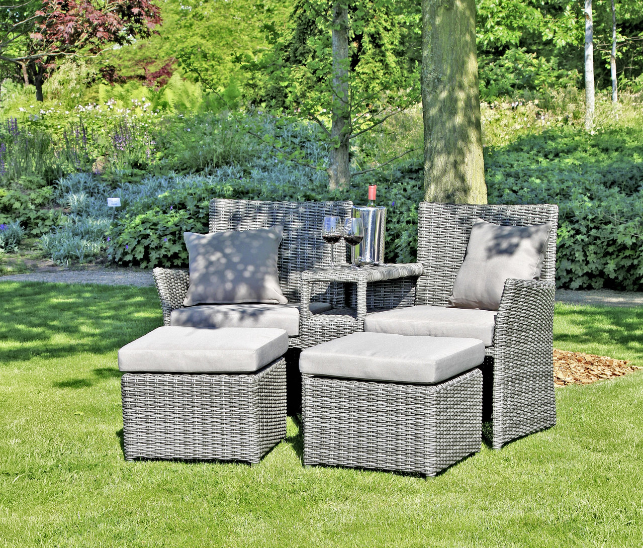 Gartenmöbel-Set aus grauem Rattan mit Kissen, Tisch, Weinkühler, Flasche und Gläsern auf Rasen vor Bäumen.